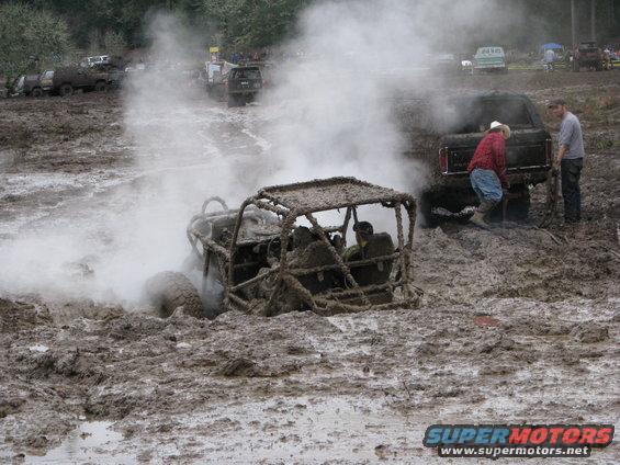 Mud-covered off-road buggy stuck in mud, emitting steam, with people and other vehicles nearby