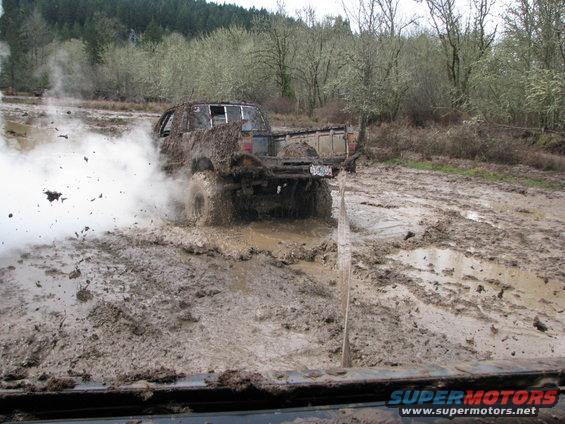 Mud-covered pickup truck spinning tires in a mud bog, kicking up dirt and smoke