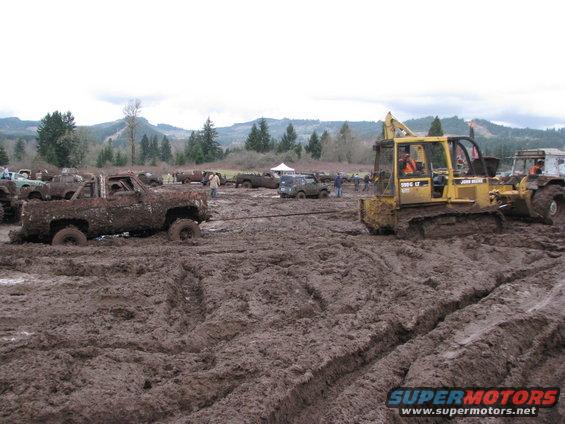 Mud-covered pickup truck being pulled by a bulldozer in a muddy field with other vehicles and trees in the background