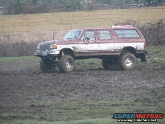 Maroon and white four-door SUV with large tires driving through mud. Appears to be a 1990s Ford Bronco or similar