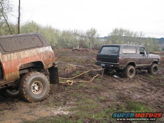 Black 1978 Ford Bronco being towed by a mud-covered orange and white SUV in a muddy field