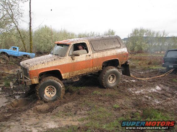 Mud-covered orange and white SUV stuck in mud, with a blue truck behind it