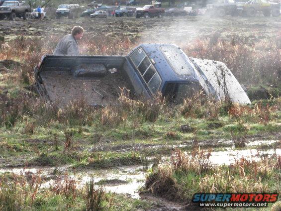 Blue pickup truck stuck in mud with a person in the bed. Other vehicles visible in background