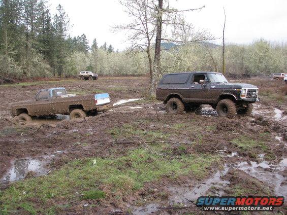 Muddy brown 1978 Ford Bronco navigating a muddy field with a pickup truck stuck nearby