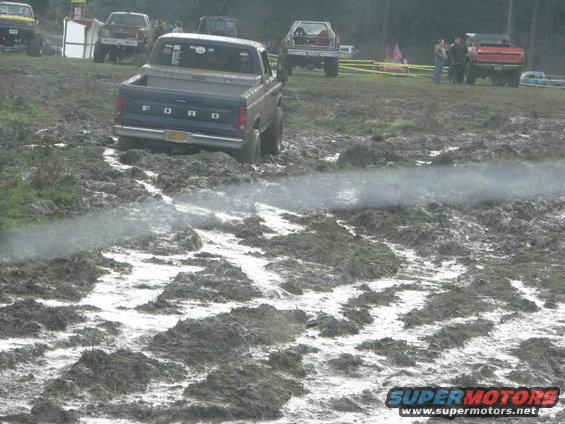 Blue Ford pickup truck driving through mud; other trucks visible in background