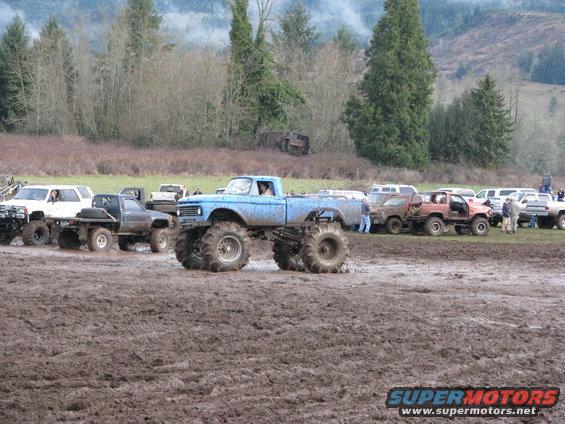 Blue monster truck with large tires in a muddy field, other trucks and people in background