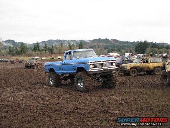 Blue lifted pickup truck with large tires in mud, other vehicles in background