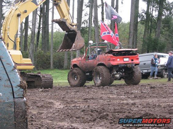 Red off-road vehicle with large tires in mud, excavator in background, van and people to the right