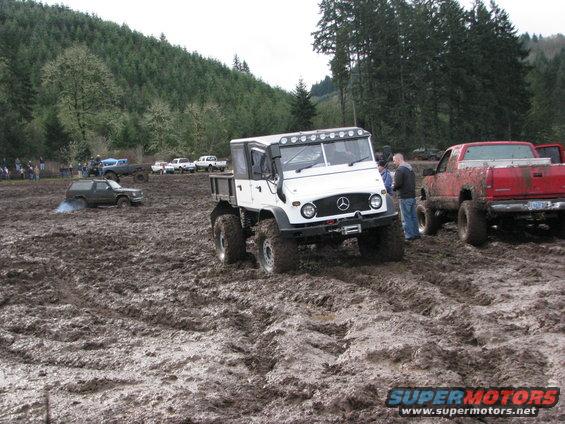 White Mercedes-Benz Unimog with large tires in mud, red pickup truck nearby