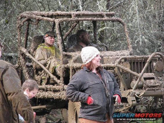 Muddy off-road vehicle with people inside and standing around it in a wooded area