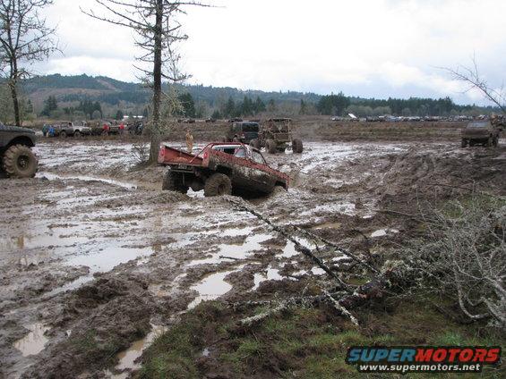 Several off-road trucks stuck in deep mud during an event, with trees and hills in the background