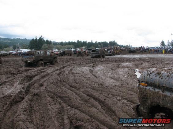 Muddy trucks and SUVs parked and driving in a large mud pit with trees in the background