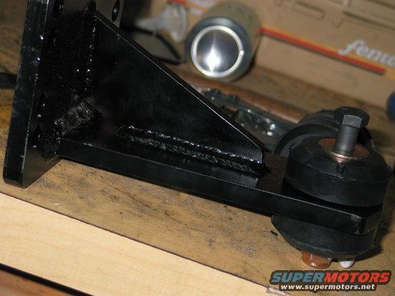 Black metal bracket with rubber mounts and a bolt, resting on wood. A can and box are in the background