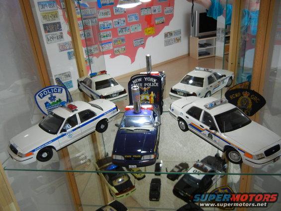 Collection of police car models, including a New York State Police car and others with police markings, displayed on glass shelves