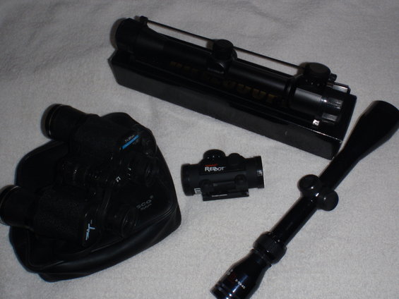 Black rifle scope, red dot sight, and binoculars on a white surface