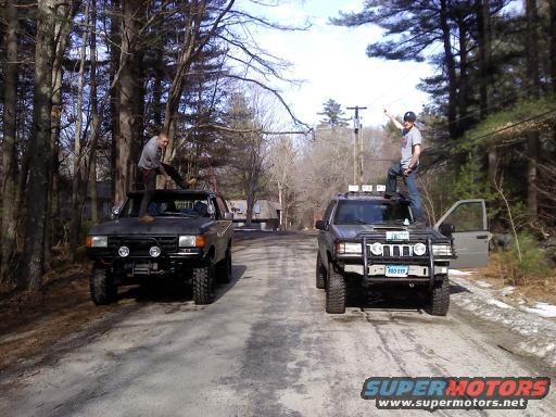 Two modified SUVs, one black and one gray, parked on a dirt road with people standing on their roofs. The gray SUV has a license plate reading '980 EXP'