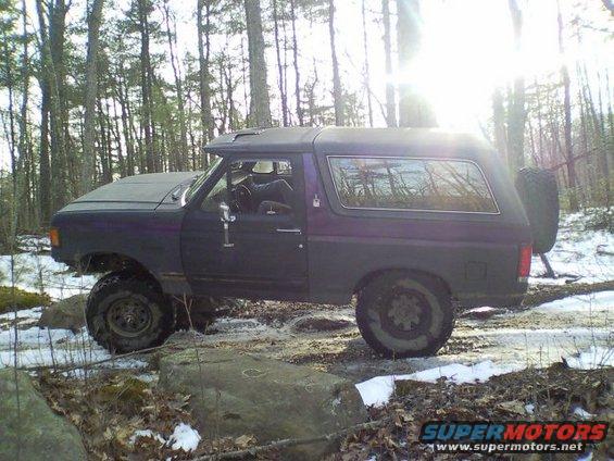 1989 Ford Bronco parked on rocky terrain with snow on the ground, surrounded by trees
