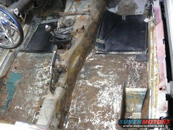 Car interior floor with rust and exposed metal, showing a manual transmission shifter and part of a steering wheel