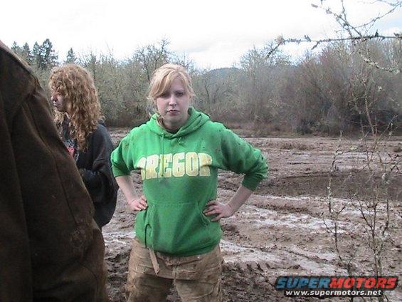 Woman in green "Oregon" hoodie and muddy pants stands in a muddy field