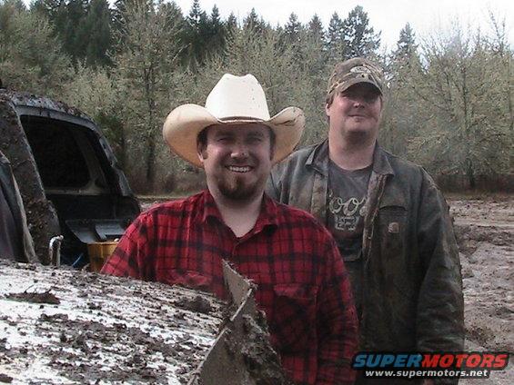 Two men, one in a cowboy hat, stand in a muddy area with trees in the background
