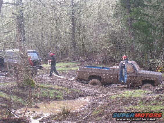 Two pickup trucks stuck deep in mud in a forest, one being pulled by the other