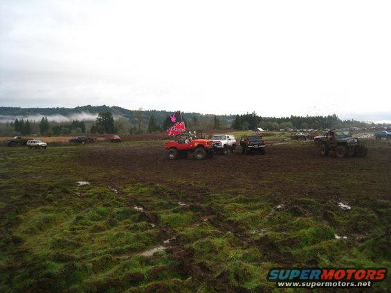 Red off-road vehicle with flags flying in a muddy field with other vehicles