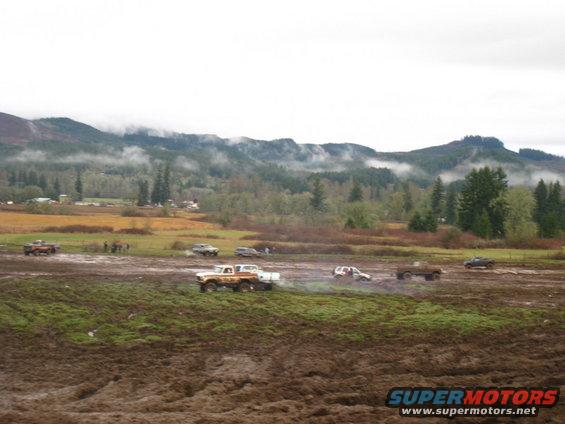 Several trucks and SUVs driving through a muddy, grassy field with trees and mountains in the background