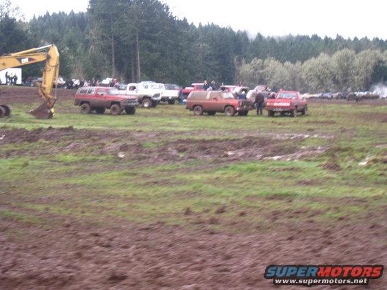Muddy field with several off-road vehicles, including red Broncos and white trucks, gathered near trees. An excavator is on the left