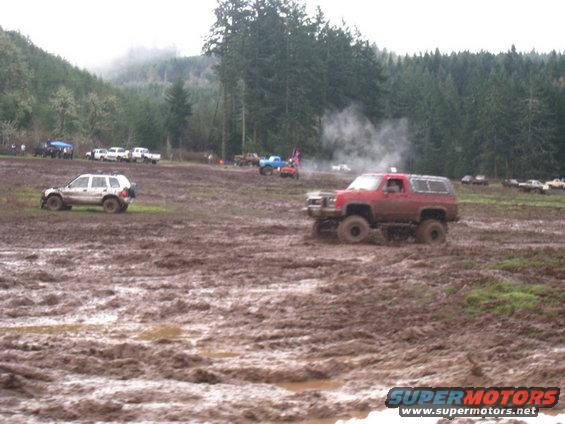 Red SUV driving through a muddy field with trees in the background