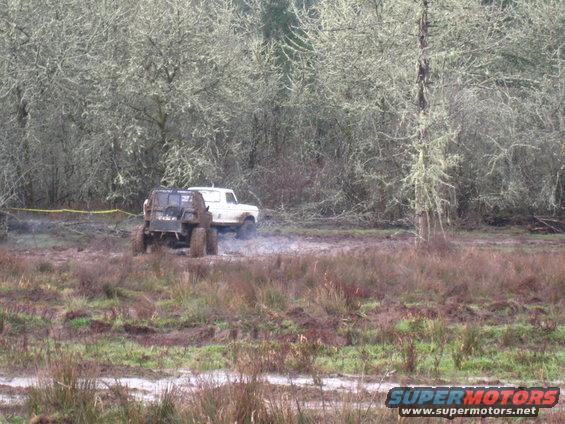 White Ford Bronco and a dark ATV stuck in mud, trees in background