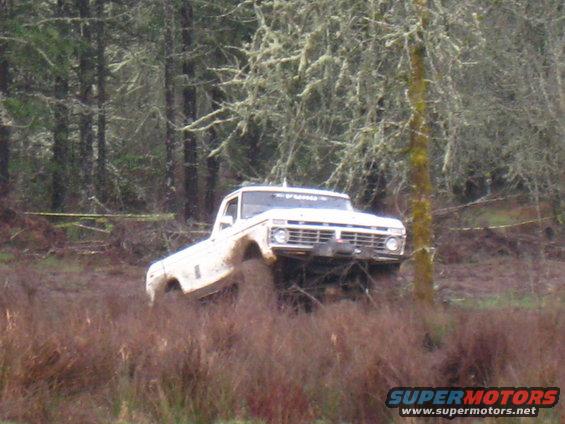 White pickup truck with mud splashing, driving through a muddy field with trees in the background