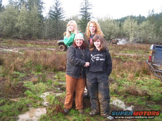 Four people stand in a muddy field with trees in the background. Two off-road vehicles are visible