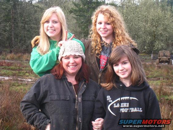 Four people pose in a muddy outdoor area with a vehicle in the background