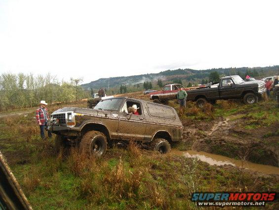 Mud-covered 1978 Ford Bronco with a person in a cowboy hat driving, in a muddy field with other trucks