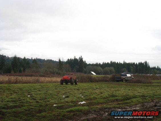 Red off-road buggy and a pickup truck in a muddy field with trees in the background