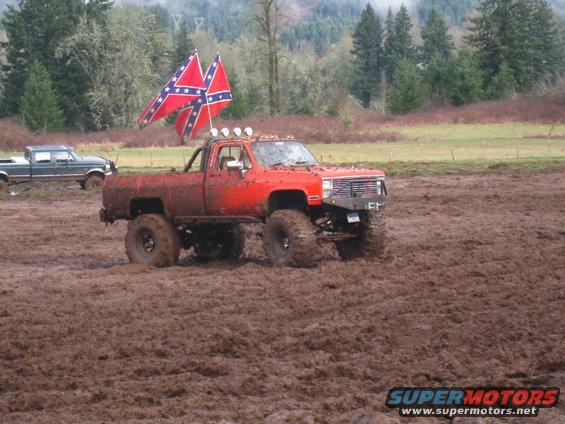 Red lifted pickup truck with two confederate flags flying, stuck in mud. Another dark pickup is in the background