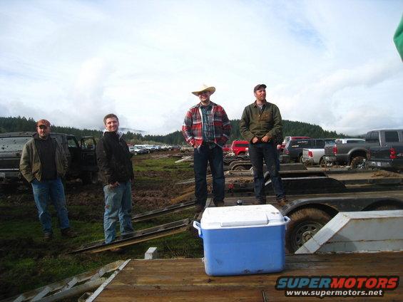 Four men on a trailer with trucks in muddy field