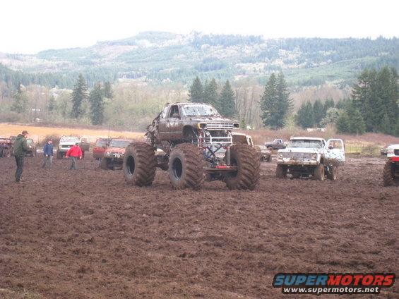 Monster truck with large tires sits in mud, other trucks and people in background
