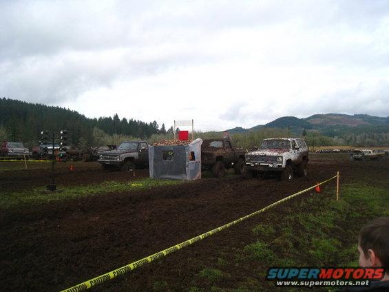 Muddy field with several trucks, including a white SUV with red mud splatters, and a grey pickup truck