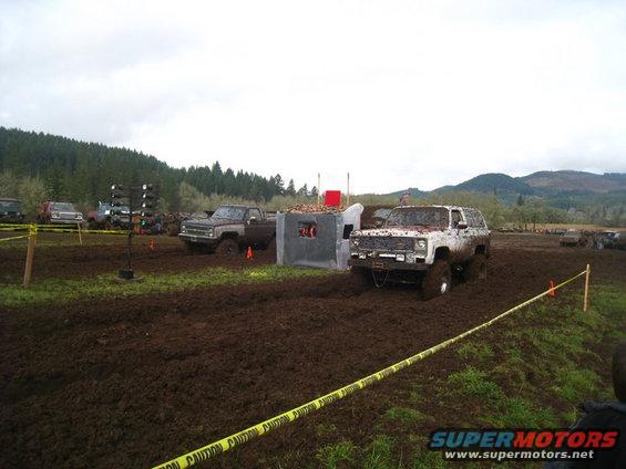 White SUV stuck in mud, front view. Other vehicles and muddy terrain visible