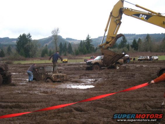 Mud bog event with several vehicles stuck in mud, an excavator lifting mud, and people watching