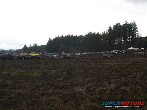 Muddy field with numerous trucks and SUVs parked and driving, trees in background
