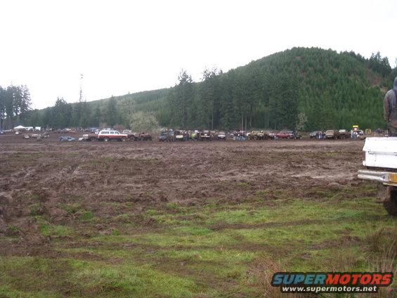 Muddy field with many trucks and SUVs parked in the distance. A person stands on the back of a white truck in the foreground