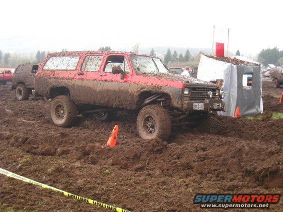 Red SUV covered in mud, driving through a mud pit with orange cones and a tent structure nearby