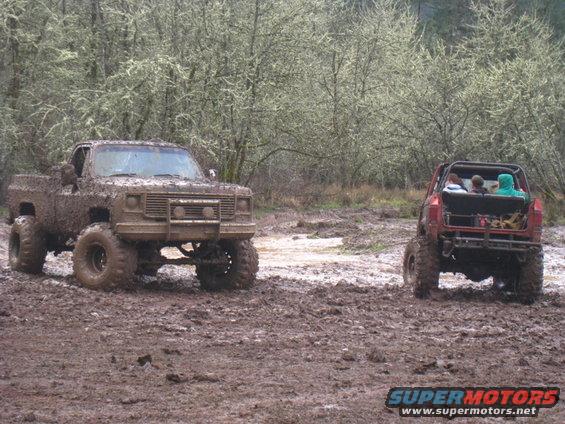 Mud-covered pickup truck and red off-road vehicle in a muddy field with trees