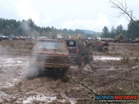 Mud-covered red van stuck in mud, with other vehicles in the background