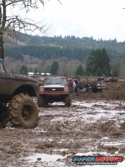 Red pickup truck with large tires in mud, front view. Other vehicles and people in background