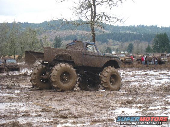 Monster truck with large tires stuck in mud, other vehicles and people in background