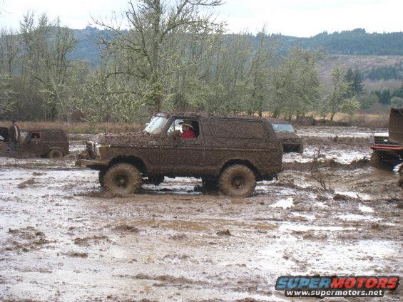 Mud-covered 1978 Ford Bronco in a muddy field with trees and hills in the background
