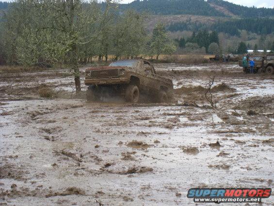 Mud-covered pickup truck stuck in a muddy field, other vehicles and people in background